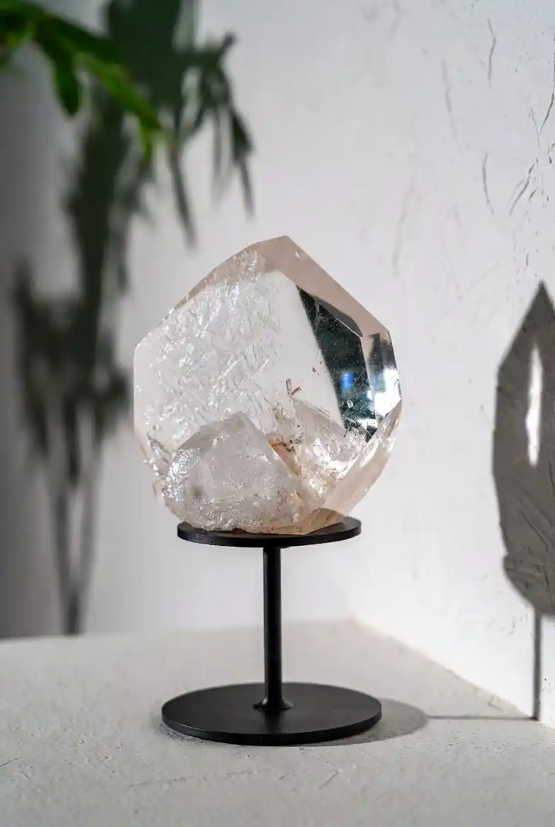 Pièce de collection unique Gemska : cristal de quartz transparent sur socle noir minimaliste, fond mur blanc texturé et marbre clair.