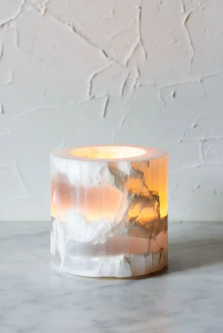 Bougeoir cylindrique en sélénite blanche Gemska allumé, effet photophore lumineux sur marbre et mur blanc texturé.